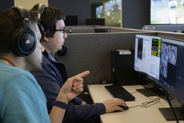 A man dressed in scrubs and wearing headphones points at a computer screen that Lucas Grijalva sits in front of and monitors. 
