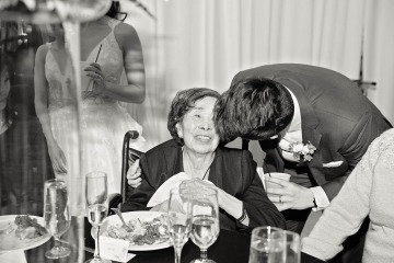 A black and white image of a man at a wedding reception leaning over to hug his grandmother who is in a wheelchair