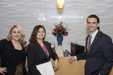 Anesa Castillo, Virginia Zazueta-Blackman and Ross Dubois stand in front of a vase of red roses with the UArizona College of Medicine – Tucson logo behind them on the wall. 