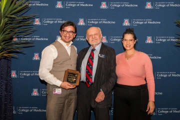 Two College of Medicine – Tucson faculty members and a donor pose for a photo in front of a college-logoed backdrop. One is holding an award.
