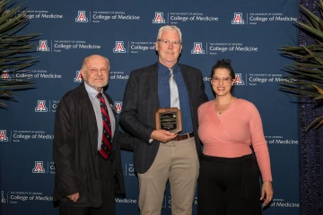 Two College of Medicine – Tucson faculty members and a donor pose for a photo in front of a college-logoed backdrop. One is holding an award.
