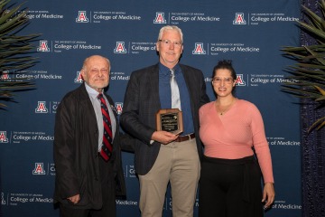 Two College of Medicine – Tucson faculty members and a donor pose for a photo in front of a college-logoed backdrop. One is holding an award. 