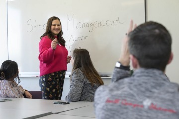 Anna Alkozei stands at the front of class with medical residents seated at a table in front of her. A whiteboard behind her reads “Stress management.” 