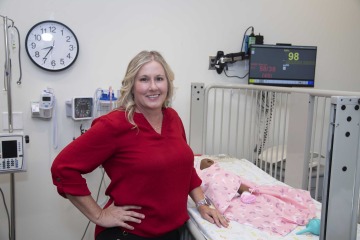 Lecturer Kelleigh Beatty stands next to a simulated baby in a bed.
