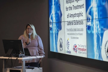 a student standing at a lectern holding a microphone in front of a projection screen showing a research project title page