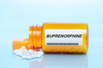 prescription medication bottle labeled “buprenorphine” lays on its side with white pills spilling out on a light blue background