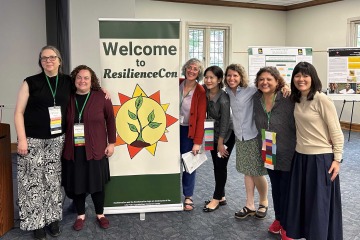 Seven woman stand in a conference room surrounding a banner that says “Welcome to ResilienceCon.”