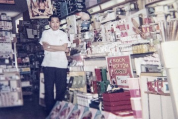 A man in a white shirt stands in front of displays in his pharmacy. The photo is several decades old.
