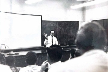 Albert L. Picchioni stands in front of a blackboard in a classroom full of people holding a book in a black and white photo.
