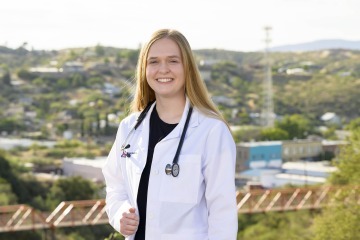 Portrait of Primary Care Physician Scholarship recipient Emma Rautenberg wearing a doctor’s white coat and stethoscope with a rural Arizona town in the background.
