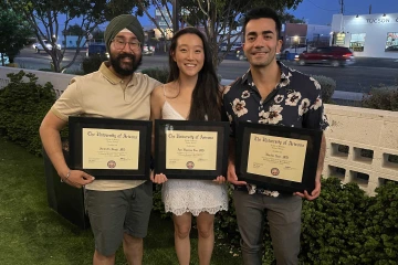 Three doctors in casual clothes holding plaques during a fellowship graduation ceremony on a restaurant patio