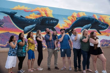 A group of people in front of a mural of whales swimming in the desert.  