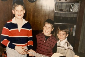 An old photo of three boys standing in a row holding a guitar.