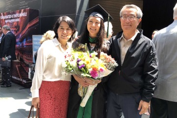 Woman wearing graduation cap and gown and holding flowers poses with her parents