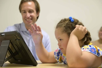 Dan Combs is smiling in the background while a young girl looks at a computer screen. 