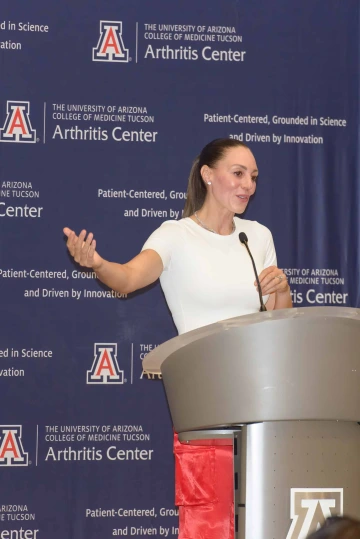 University of Arizona women’s basketball coach Adia Barnes speaks at a lectern in front of a blue backdrop with U of A Arthritis Center logos on it.