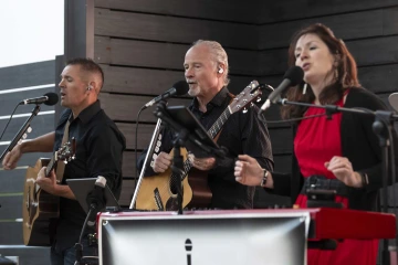 Photo of two men singing and holding guitars in front of microphones next to a woman singing into a microphone.