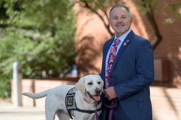 Professor Jim Reed stands outside wearing a suit and tie with his golden lab, Jenny. 