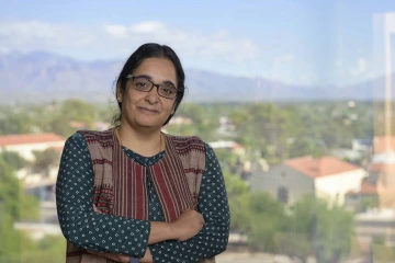 Portrait of physician-scientist Dr. Lalitha Madhavan, standing in front of a window overlooking Tucson with mountains in background.