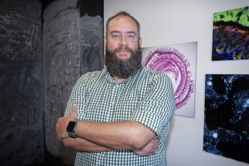 Portrait of a man in a checked shirt standing with his arms crossed in front of artwork.