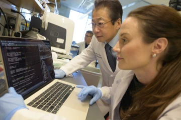A man in glasses and a lab coat looks over the shoulder of a woman in a lab coat and gloves as they examine information on a laptop.