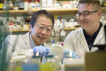 Two men in white lab coats and glasses closely examine a petri dish while surrounded by bottles of various sizes.