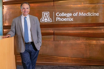 A man in a suit coat stands in front of a wall that has College of Medicine – Phoenix lettering.