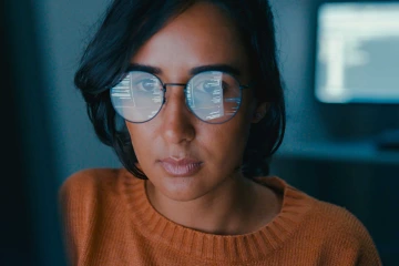 A photo illustration of a woman looking at a computer screen with words and numbers reflected in her glasses.