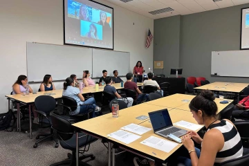 Gabriela Valdez, PhD, stands in front of students in a classroom and people on a screen.