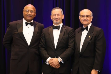 Three men in black tuxedos smile as they stand next to each other in front of a blue curtain background. 