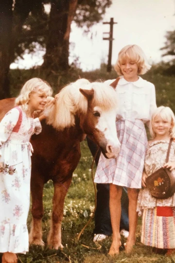 A family photo of young Kacey Ernst in a long dress standing next to a pony with her two sisters.