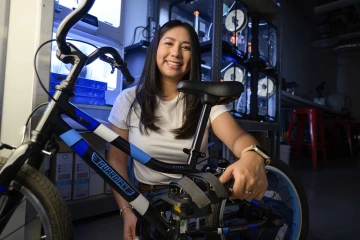 A Health Sciences Design student kneels down with a bicycle equipped with an adaptive foot pedal she designed