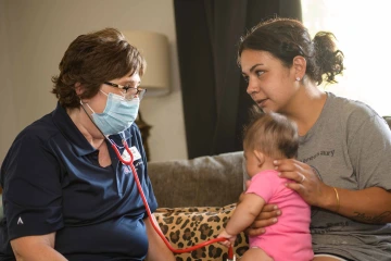 A woman wearing a facemask and stethoscope speaks with a mother with a baby on her lap