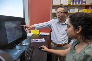 Mingyu Liang, MB, PhD, next to a computer with his finger pointing out something on the monitor. Lab manager Bhavika Theranialso stands in front of the monitor, staring at what he’s pointing at.