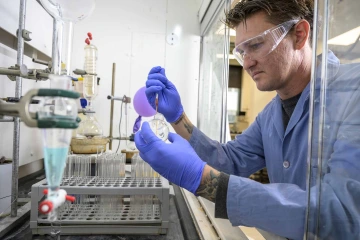 A person wearing a blue lab coat and gloves is working in a research lab, pipetting a red liquid mixture into a glass tube, surrounded by lab equipment