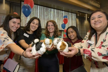 A group of students stands in front of red and blue balloons holding stuffed guinea pig toys.