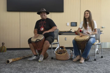 A man wearing a hat smiles as he pounds a drum tucked between his knees while a woman seated next to him also hits a drum.