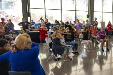A room full of people sitting in chairs hold their arms off to the side during a dance activity.