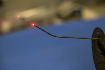 A close-up of a gloved hand holding a thin, flexible falloposcope with a red light emitting from the tip, set against a blurred lab background.