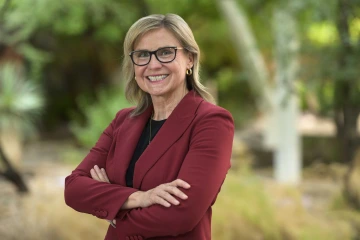 An outdoor portrait of Jennifer Barton, PhD, smiling, wearing a red blazer and glasses.