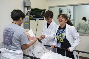 Three middle school students stand around manikin in a hospital bed. One student holds a pink bed pan up to the manikin’s mouth.