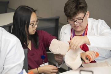 While a medical student extends her arm, a middle school student in a doctor’s coat wraps a bandage around it.