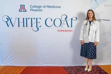 Katie Simic stands smiling in a white lab coat with a stethoscope around her neck, posing in front of a backdrop for the University of Arizona College of Medicine – Phoenix White Coat Ceremony.