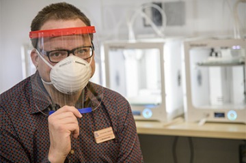 Tory Middlebrooks, of University Libraries, models a face shield, which is designed to be worn in conjunction with addition PPE such as a mask. Middlebrooks mans the 3D printers that produced the headbands for the face shield.