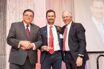 Three men in suits and ties looking at camera and smiling. Man in middle is holding a crystal award. 