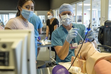professor performs a simulated clinical procedure on a mannikin while student watches monitor
