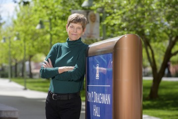 Woman with short brown hair wearing a green turtleneck and black pants leans again the University of Arizona Roy P. Drachman Hall sign