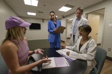 Four people in a room. Two women are sitting at a desk with one wearing a white coat and the other dressed casually in a pink hat and athletic top. Two other people, a man wearing a white coat and a woman wearing a light blue suit, look at the seated people.