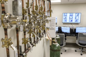 A wall of tubes hooked up to gasses in the forefront. A person is seen at a desk with a computer screen with another monitor observing sleep study participants in the background.