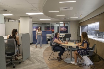 People utilize the space in a breakroom with tables and a desk.